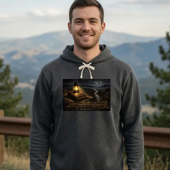 Smiling young man at a mountain overlook wearing a pepper garment-dyed hoodie printed with a painting of a brass lantern glowing on a Bible beside a night path, over a banner reading A Lamp Unto My Feet and a Light Unto My Path, Psalm 119:105